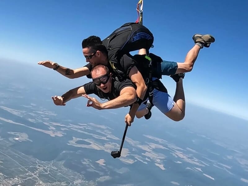 Saut en parachute en tandem avec un instructeur tenant une perche pour filmer la chute libre au-dessus des paysages.