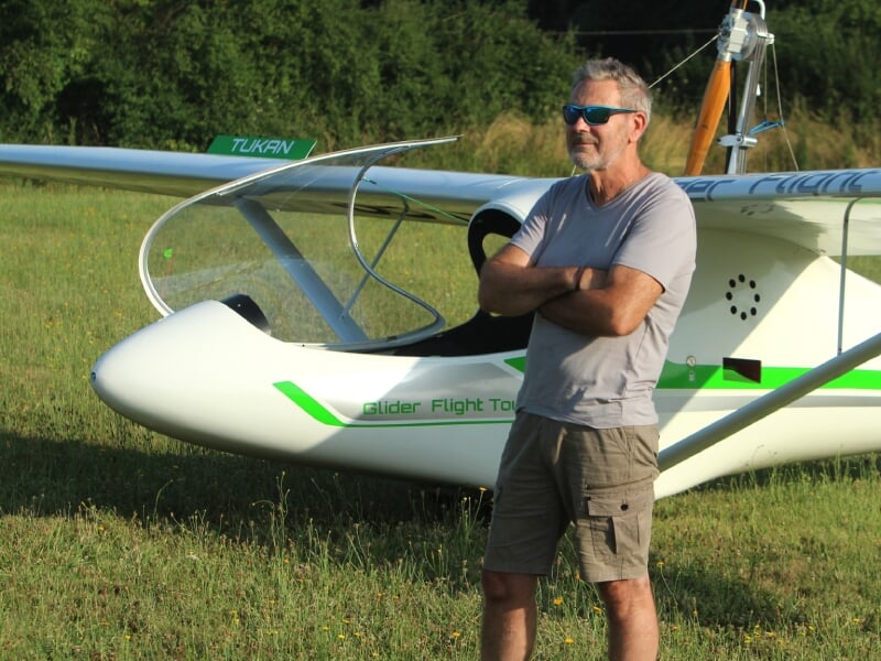 Homme aux cheveux gris et lunettes de soleil, bras croisés, se tenant devant un planeur blanc et vert marqué