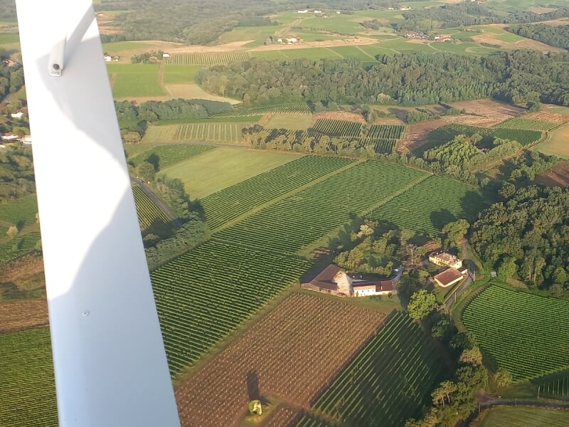 Vue depuis un planeur en vol, montrant un paysage rural composé de champs cultivés et de forêts. L'aile blanche de l'appareil est visible au premier plan, survolant une maison et des parcelles agricol