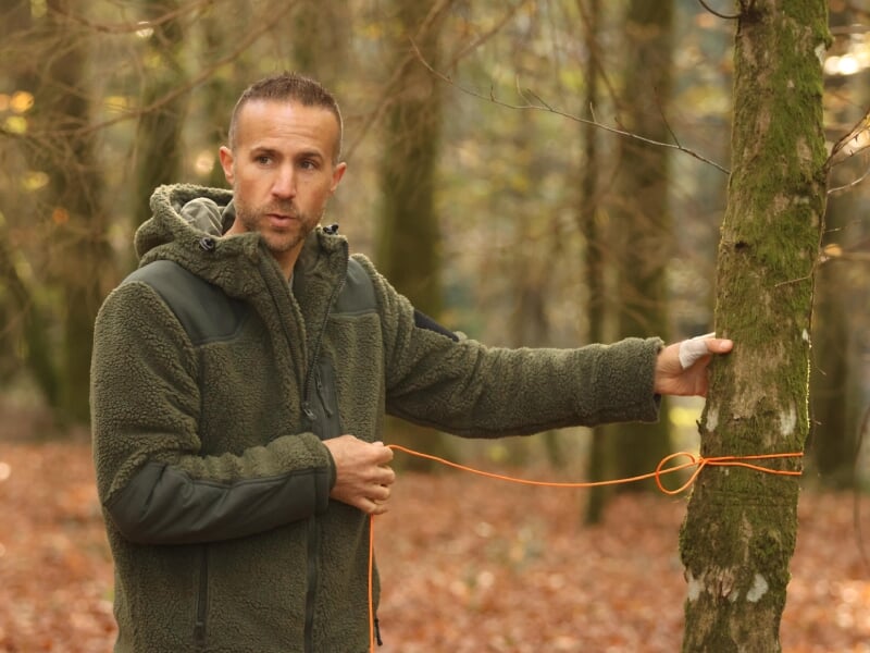 Homme en veste polaire attachant une corde à un arbre dans une forêt.