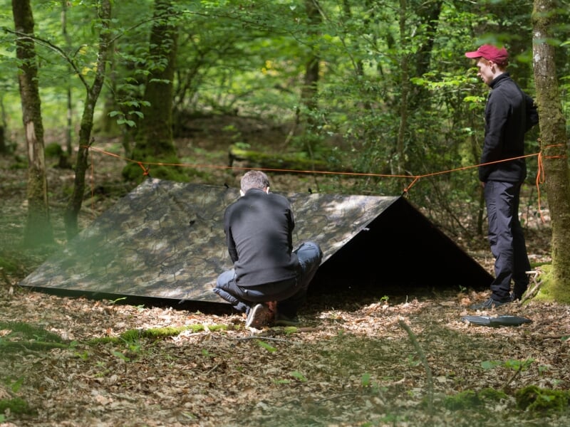 Deux hommes montent un abri de fortune en toile dans une forêt dense.