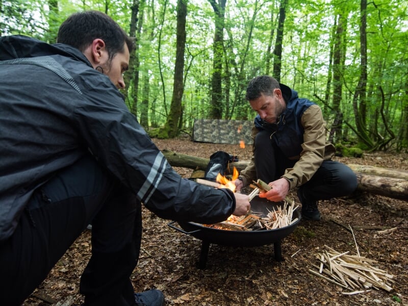 Deux hommes en tenue de randonnée allument un feu dans un brasero en pleine forêt.