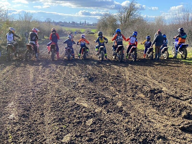 Une rangée de jeunes motards prêts à s'élancer sur un circuit en terre avec un village en arrière-plan