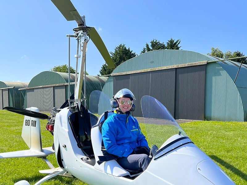 Le passager d'un autogire pose dans un appareil stationné devant les hangars d'un aérodrome de province