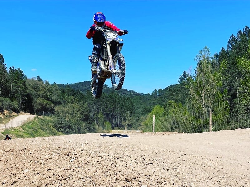 Un pilote de motocross en plein saut sur une piste en terre avec un massif forestier en arrière-plan