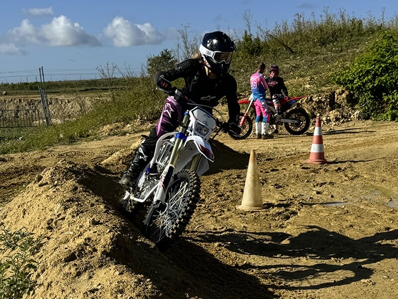 Un motard en combinaison noire et casque noir sur une moto blanche est en pleine action sur un parcours de terre, passant un obstacle tandis que deux autres motards en arrière-plan regardent, eux-même