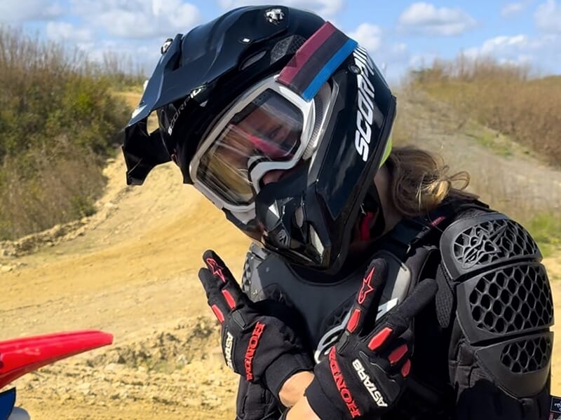 Un motard en tenue de protection noire, portant un casque noir avec des détails colorés, montre des gestes de la main tout en posant près d'une moto dans un paysage de terre et de collines sous un cie