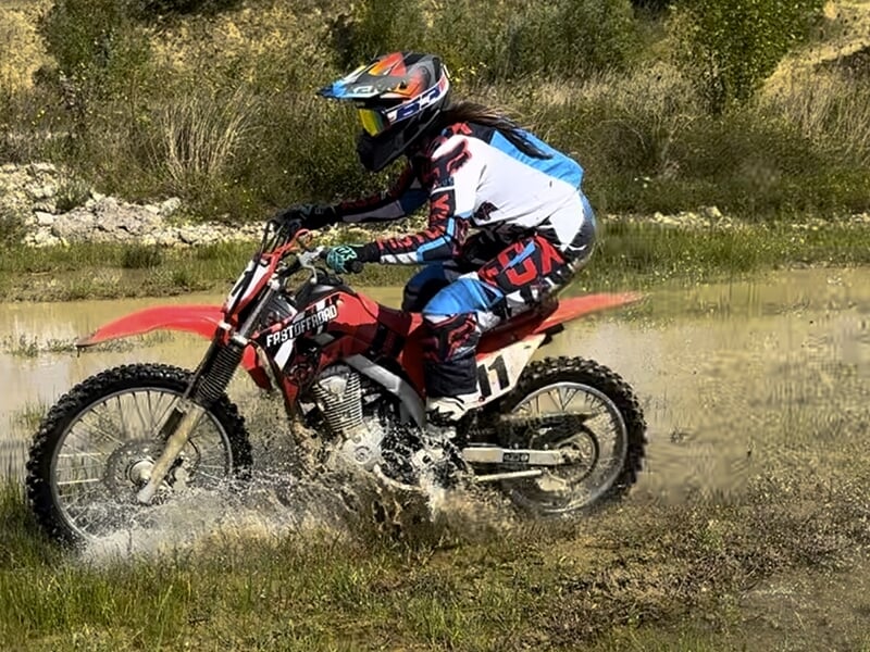 Une personne en tenue de moto-cross, avec un casque coloré et des vêtements bleus, rouges et blancs, roule sur une moto de type tout-terrain rouge à travers une flaque d'eau dans un environnement exté