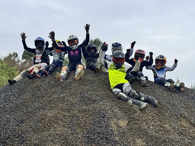 Groupe de jeunes motards en tenue complète assis au sommet d'une colline de gravier, bras levés et souriants.