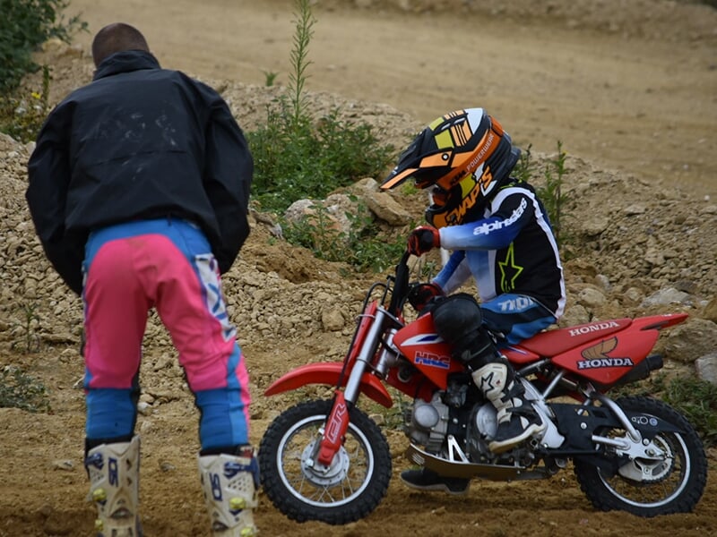 Enfant sur une petite moto tout-terrain rouge recevant des conseils d'un adulte en tenue de motocross sur un terrain de course.