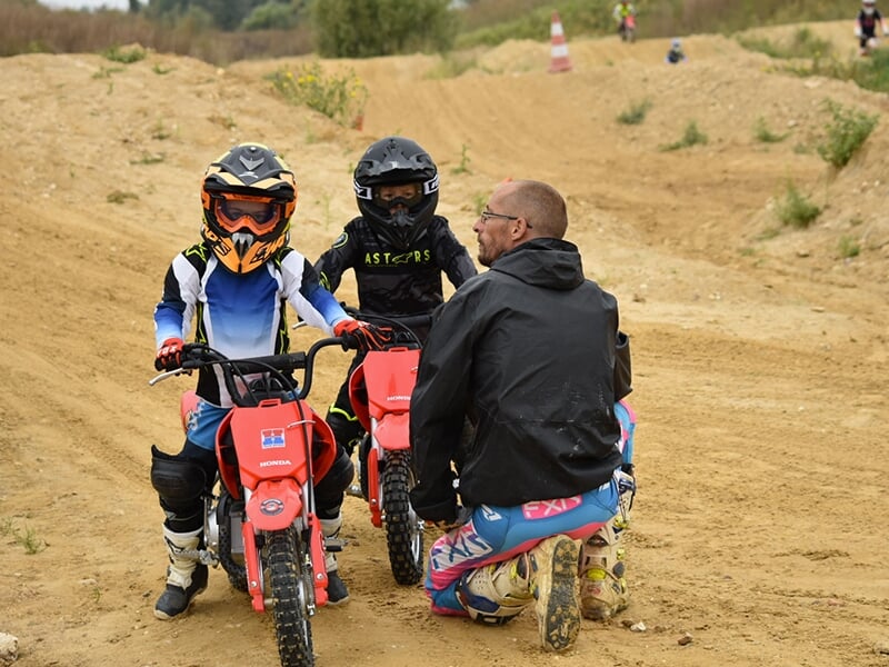 Deux jeunes motards discutant avec un instructeur sur un terrain de motocross, prêts à partir avec leurs mini motos Honda.