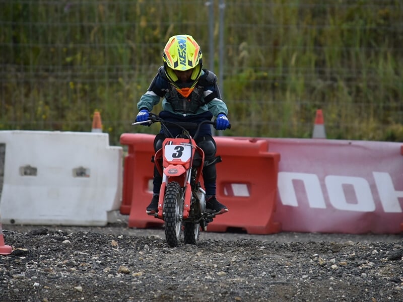  Enfant en équipement de moto pilotant une mini moto sur un terrain accidenté en pleine journée.