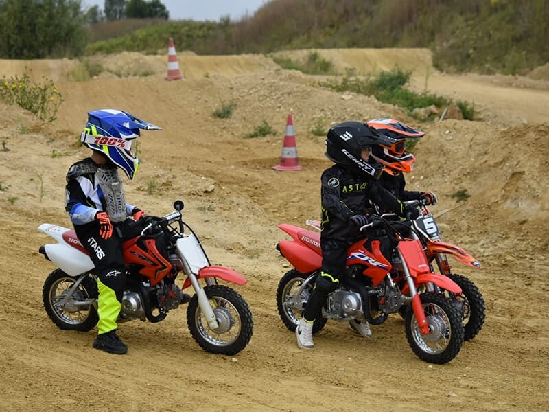 Trois jeunes motards sur des mini motos tout-terrain, discutant et se préparant à rouler sur une piste de motocross.