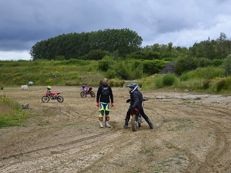Deux personnes dans un environnement de moto-cross en plein air. L'une est debout avec des vêtements colorés de protection, et l'autre est assise sur une moto. En arrière-plan, deux autres motos sont