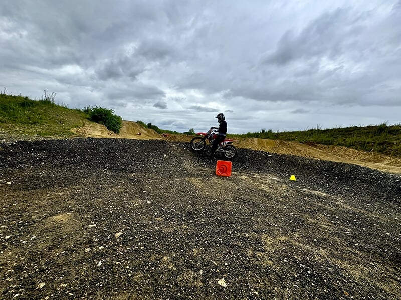 Un motard seul sur une moto rouge, s'arrêtant sur un chemin de terre noire avec des cônes et des marqueurs d'entraînement orange. Le paysage est vallonné et sous un ciel nuageux.