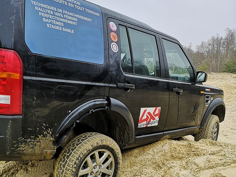 Véhicule 4x4 noir en terrain sablonneux, avec autocollants publicitaires et boue sur les roues et la carrosserie.
