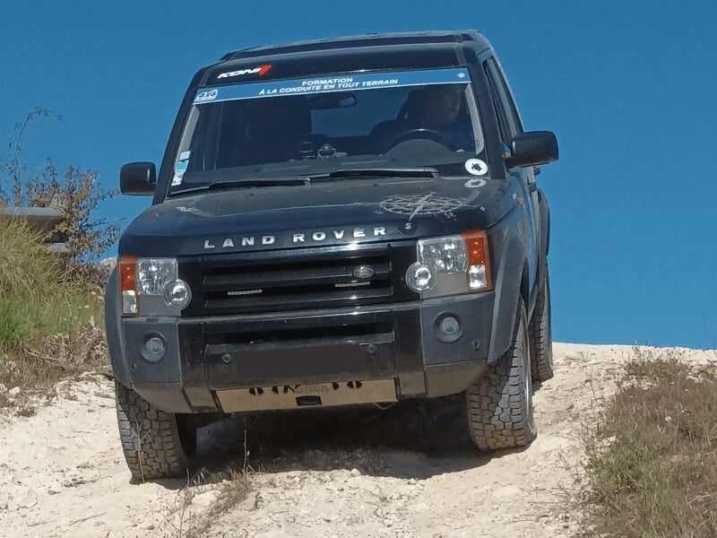 Un Land Rover noir descend une pente sablonneuse avec un ciel bleu lors d'un stage 4x4.