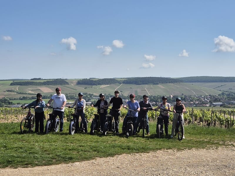 Groupe de huit personnes casquées posant sur des vélos électriques devant des vignes verdoyantes et collines sous ciel bleu parsemé de nuages blancs