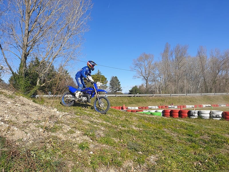 Un motocycliste descendant une pente en moto bleue, avec des pneus de couleur rouge, verte et blanche disposés autour du terrain sous un ciel bleu.