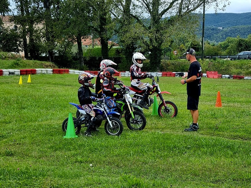  Enfants sur des mini-motos écoutant les instructions d'un entraîneur sur un circuit de motocross en plein air.