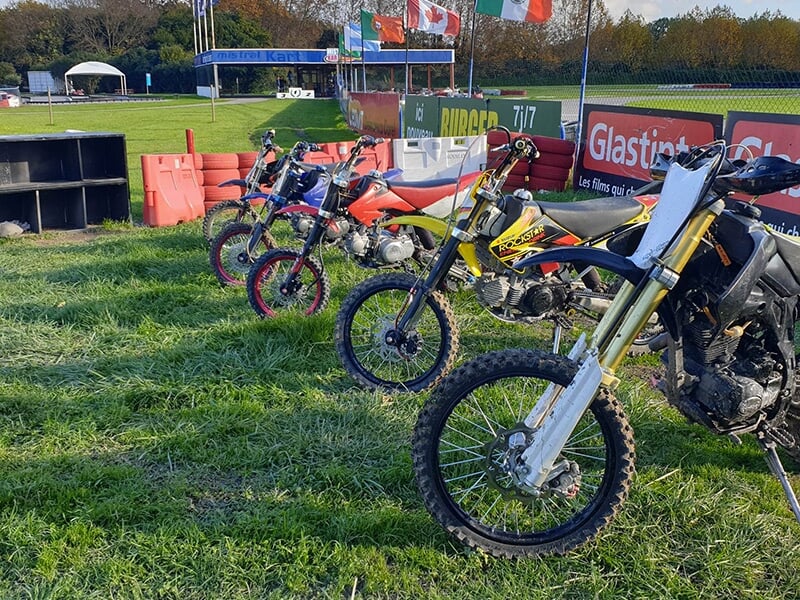 Plusieurs mini-motos garées en ligne sur de l'herbe à côté d'une piste de karting, avec des drapeaux de différents pays en arrière-plan.