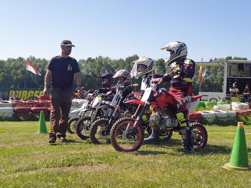 Jeunes enfants en tenue de motocross et casques, alignés sur des petites motos sur une piste d'herbe avant une course.