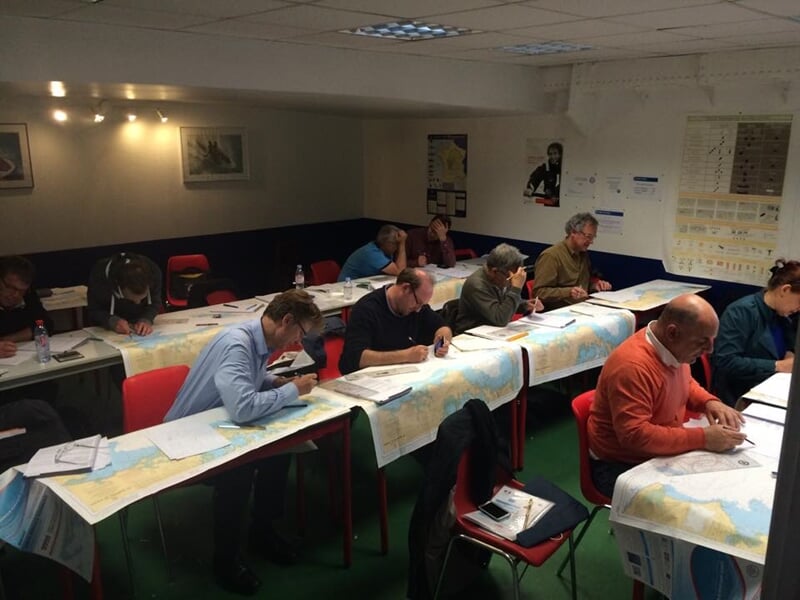 Des adultes assis en classe étudient des cartes marines sur des tables, préparant un examen de permis bateau.