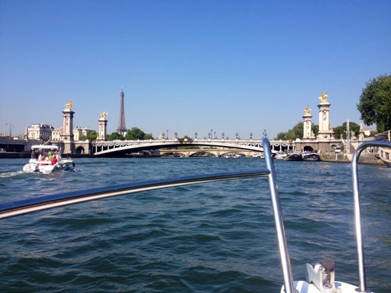 Vue depuis un bateau sur la Seine avec la Tour Eiffel et un pont historique en arrière-plan par une journée ensoleillée.