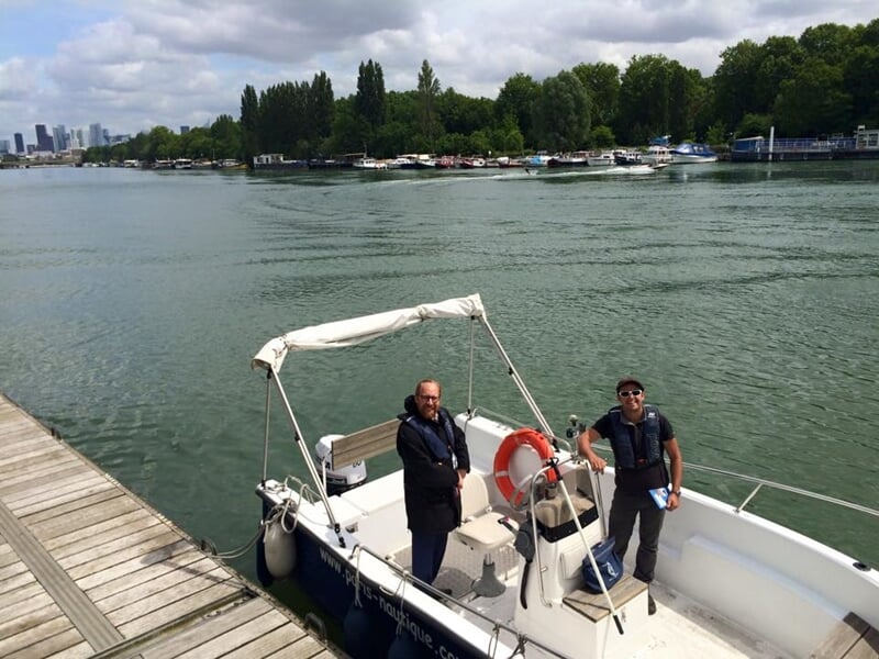 Deux hommes debout sur un petit bateau amarré au quai, avec un paysage de rivière et des arbres en arrière-plan.