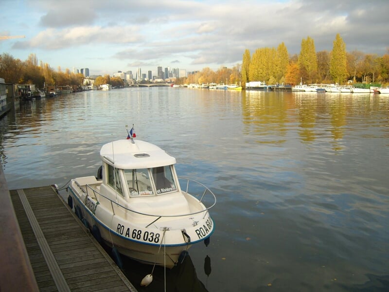 Petit bateau blanc amarré sur une rivière calme, avec des arbres aux feuilles d'automne et des bâtiments au loin.