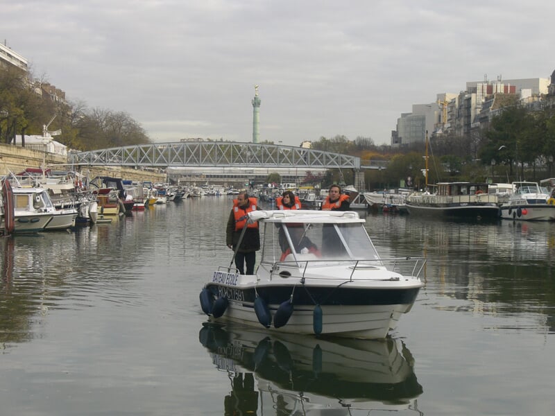 Un petit bateau à moteur navigue sur un canal urbain avec un pont métallique en arrière-plan sous un ciel nuageux.