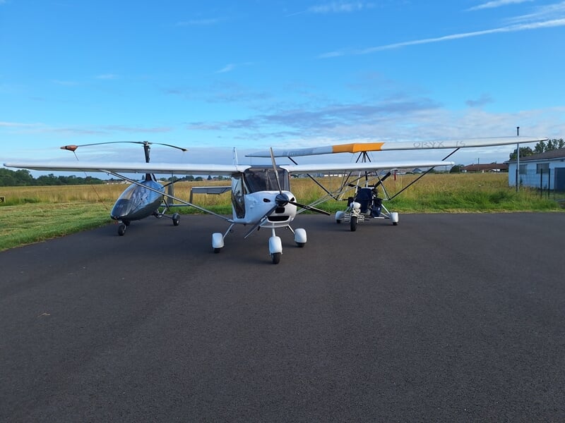 Une autogire, un multiaxes et un pendulaire ULM stationnés sur une piste d'aérodrome avec un ciel bleu et des champs en arrière-plan.