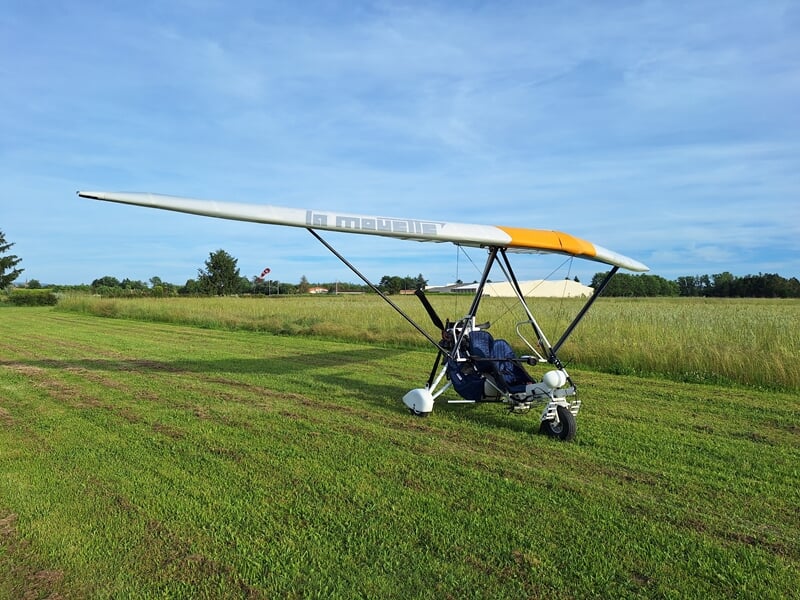 ULM pendulaire léger blanc et orange stationné sur une piste d'herbe verte sous un ciel bleu clair à l'aérodrome de Montauban.