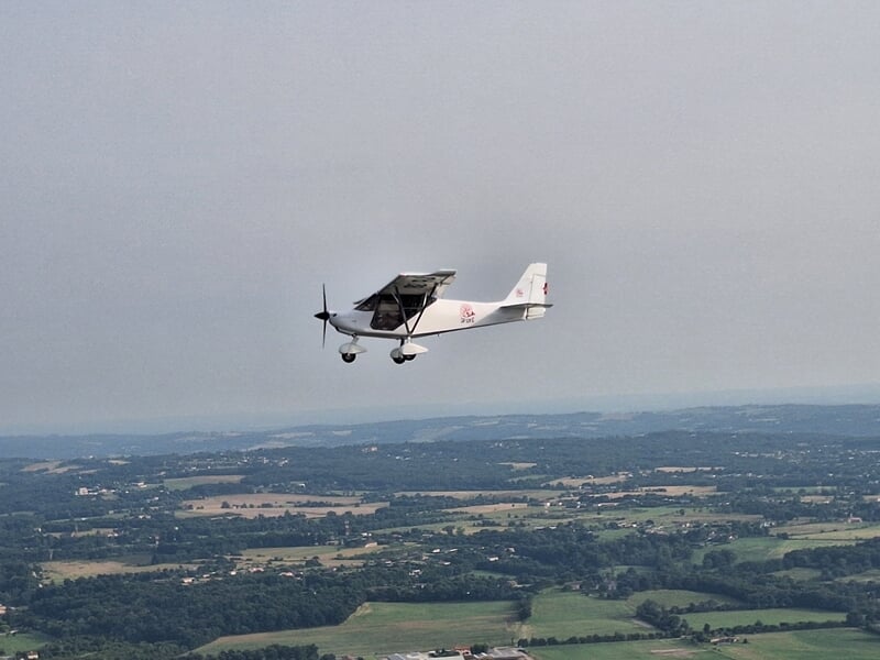 Petit avion ultraléger blanc en vol au-dessus d'un paysage rural avec champs et collines sous un ciel gris clair.
