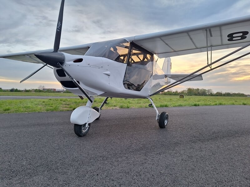 Petit avion ultraléger blanc stationné sur une piste d'aérodrome au coucher du soleil, ciel nuageux en arrière-plan.
