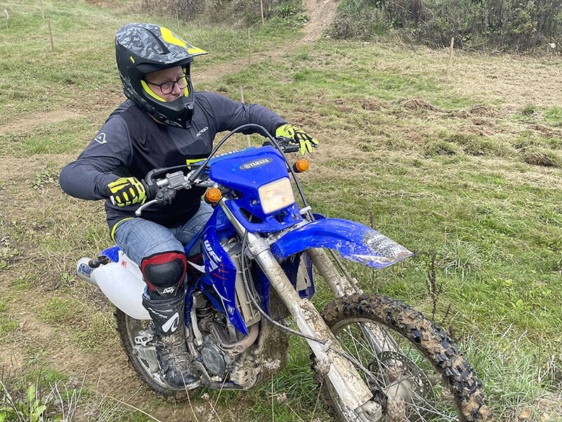 Un homme portant un casque et des lunettes de protection pilote une moto de type enduro bleue sur un terrain boueux. Il est vêtu de gants jaunes et de genouillères, concentré sur la conduite.