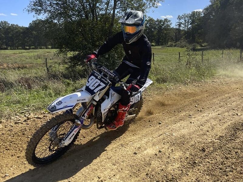 Un motocycliste en tenue noire avec des bottes rouges et un casque de protection, roulant à vive allure sur un chemin de terre. Il est sur une moto de course blanche et porte le numéro 907 sur la plaq