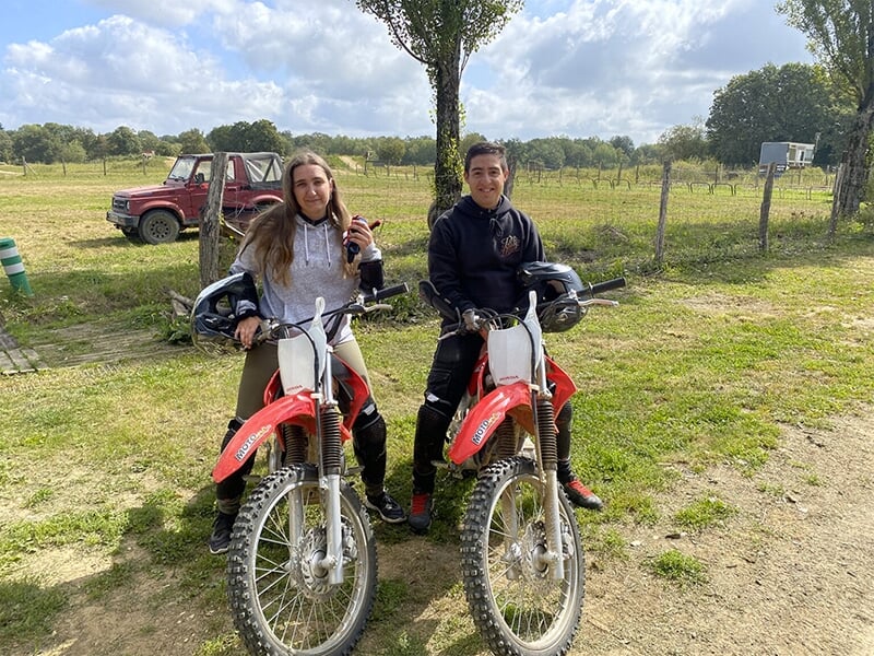 Deux jeunes, un homme et une femme, assis sur des motos de type trail rouge, souriants et prêts pour une session. Ils sont en tenue décontractée avec des casques posés sur les motos.