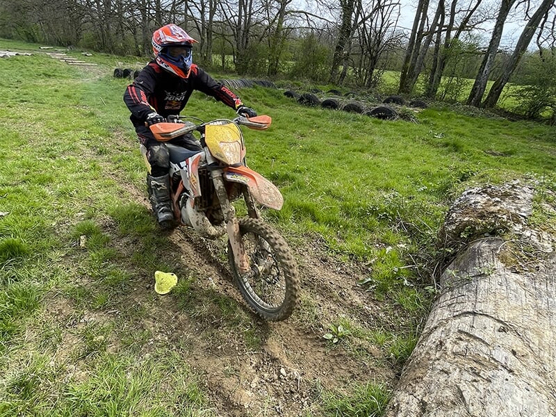 Un motard portant un casque rouge et bleu et des protections noires, franchissant un chemin boueux avec une moto de couleur orange, sur un terrain herbeux avec des pneus en arrière-plan.