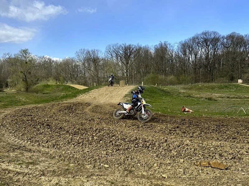 Un motocycliste en combinaison noire et casque roule sur une piste de terre dans un circuit extérieur, prenant un virage. Un autre motard est visible à l'arrière-plan sur une piste de saut.