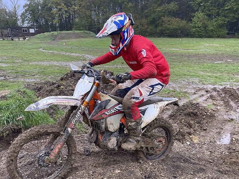Un homme vêtu d’un maillot rouge et d’un casque de protection roule sur une moto blanche et orange, traversant une piste de boue en extérieur. Sa moto est recouverte de terre, témoignant de l'état dif