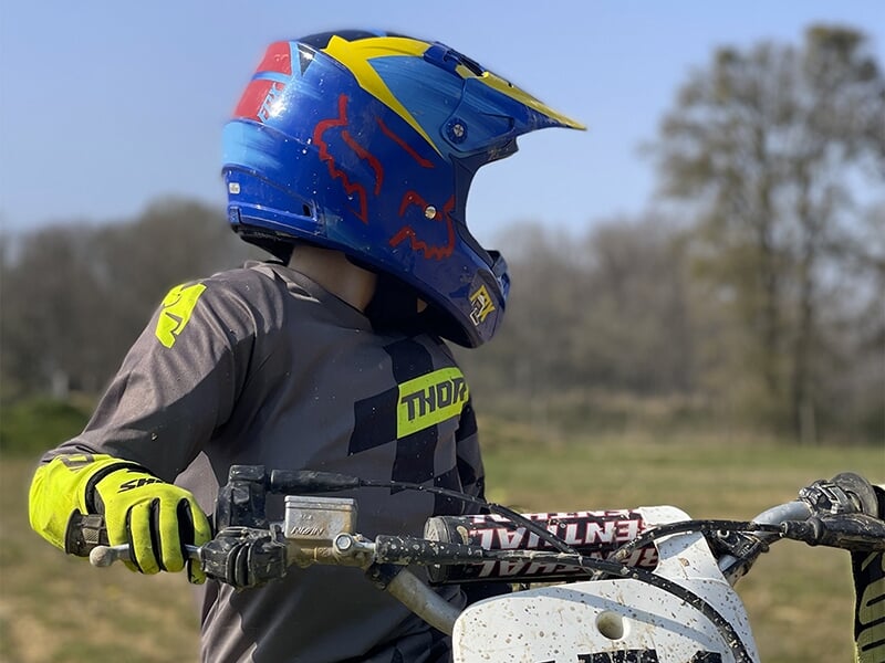 Enfant portant un casque bleu vif avec des motifs rouges et jaunes, vêtu d'un équipement de motocross Thor, assis sur une moto tout-terrain.