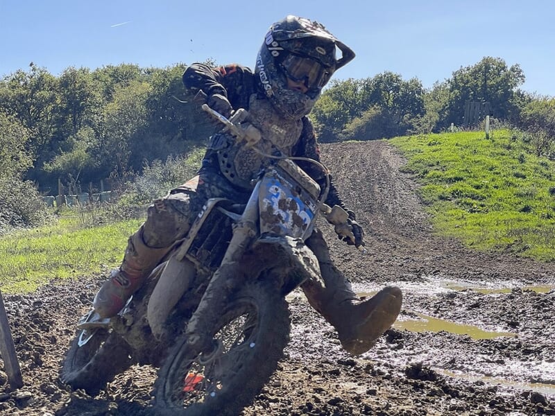 Motocycliste en pleine action sur une piste boueuse, incliné dans un virage serré avec de la boue éclaboussant autour.