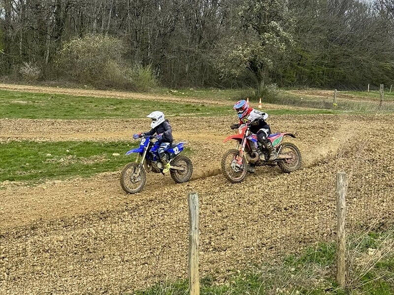 Deux jeunes motards en compétition sur une piste de terre, l'un sur une moto bleue, l'autre sur une moto rouge, se poursuivant.