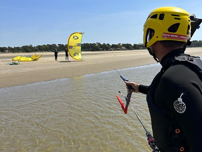 Homme portant un casque jaune tenant un cerf-volant de kite sur une plage, d'autres personnes se tenant plus loin sur le sable avec des voiles jaunes au sol.