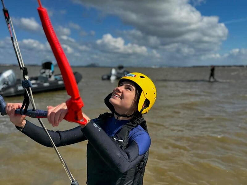Femme en combinaison et casque jaune tenant une barre de contrôle de kitesurf, regardant le ciel avec le sourire sous un ciel nuageux.