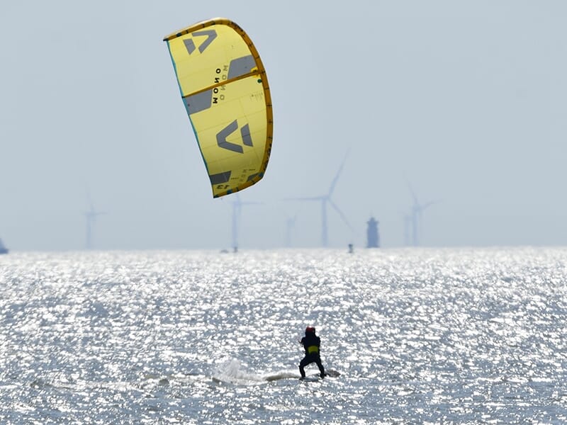 Kitesurfer glissant sur l'eau avec une voile de kite jaune, des éoliennes visibles à l'horizon sous un ciel gris.