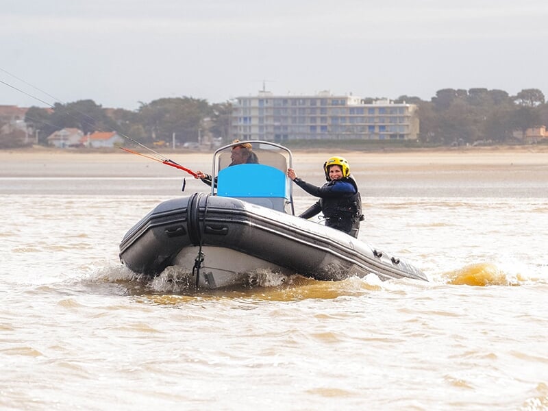 Bateau pneumatique avec deux personnes à bord, l'une d'elles portant un casque jaune et tenant un équipement de kitesurf, plage en arrière-plan avec quelques bâtiments.