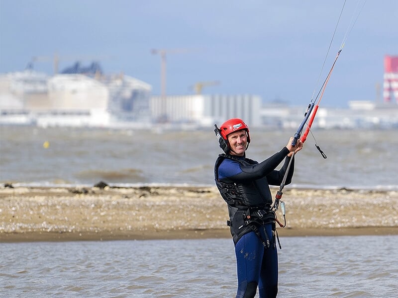 Homme souriant en combinaison et casque rouge sur une plage, tenant une barre de contrôle de kitesurf avec un arrière-plan industriel flou.