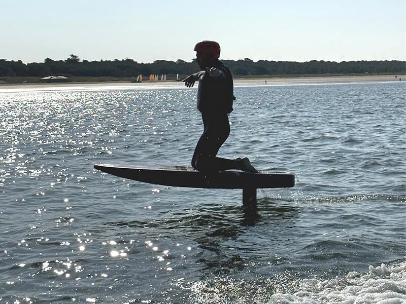 Personne en combinaison et casque, sur une planche e-foil au-dessus de l'eau, au lever ou coucher du soleil. La silhouette du surfeur se détache sur l'eau scintillante, avec l'hydrofoil visible sous l